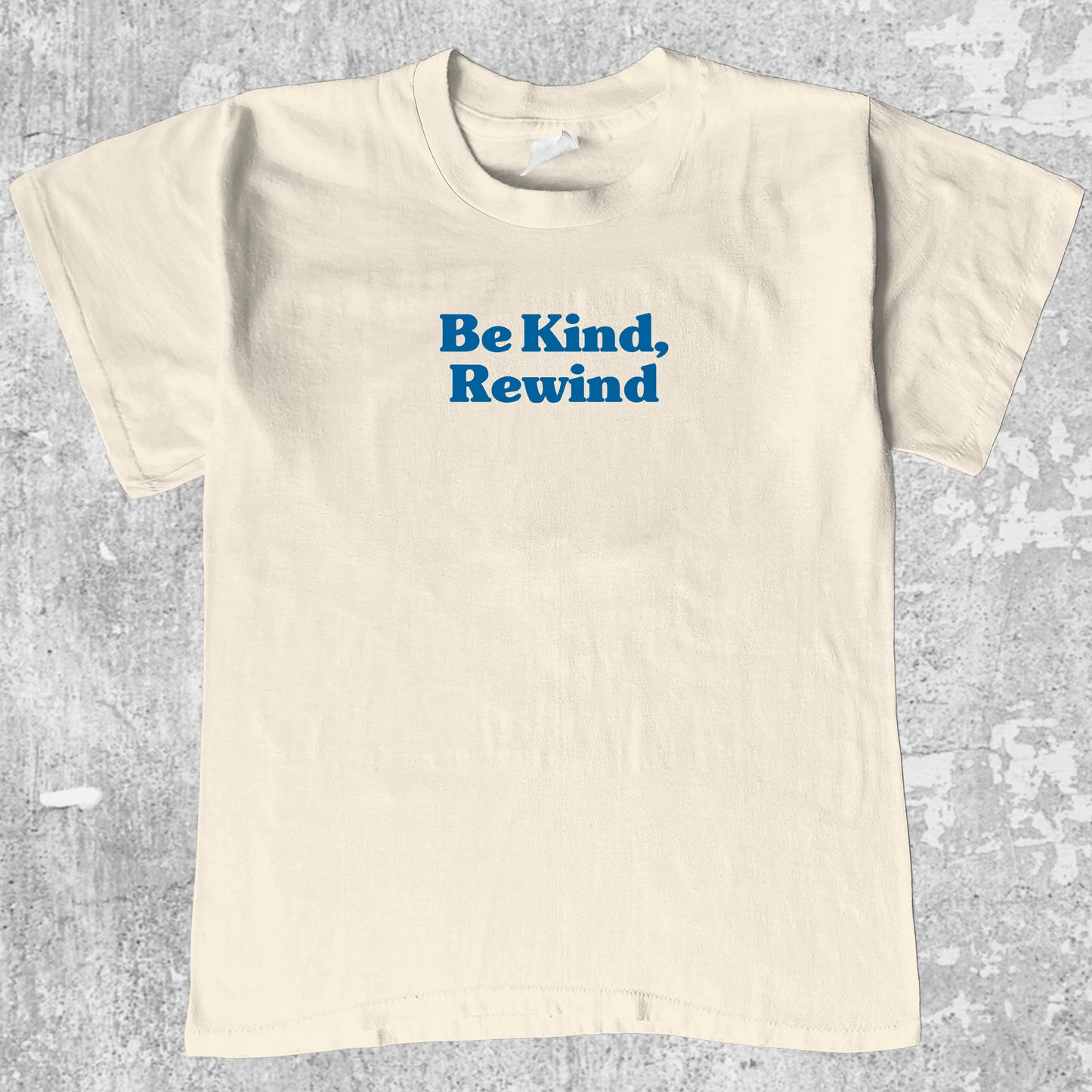 Beige t-shirt with 'Be Kind, Rewind' text on a gray textured background