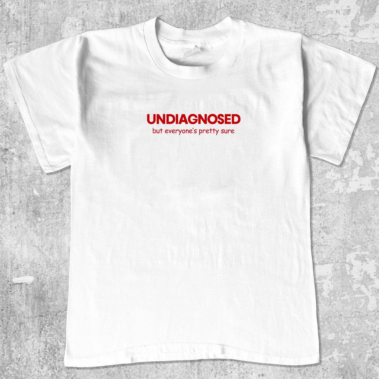 White t-shirt with red text 'UNDIAGNOSED but everyone's pretty sure' on a gray textured background