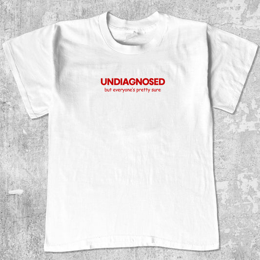 White t-shirt with red text 'UNDIAGNOSED but everyone's pretty sure' on a gray textured background