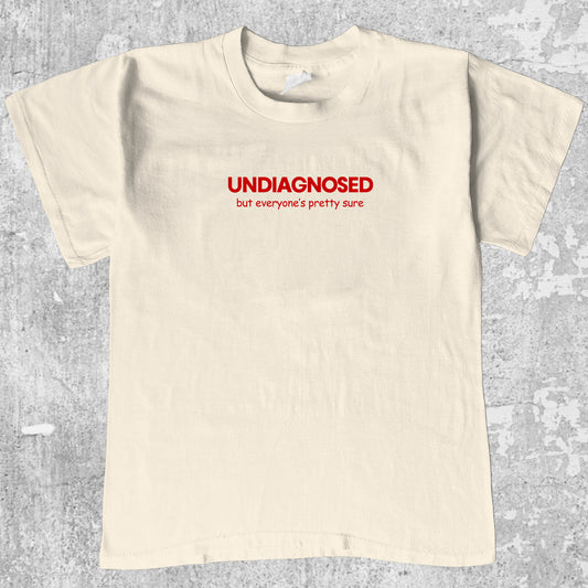 Beige t-shirt with red text 'UNDIAGNOSED but everyone's pretty sure' on a gray textured background